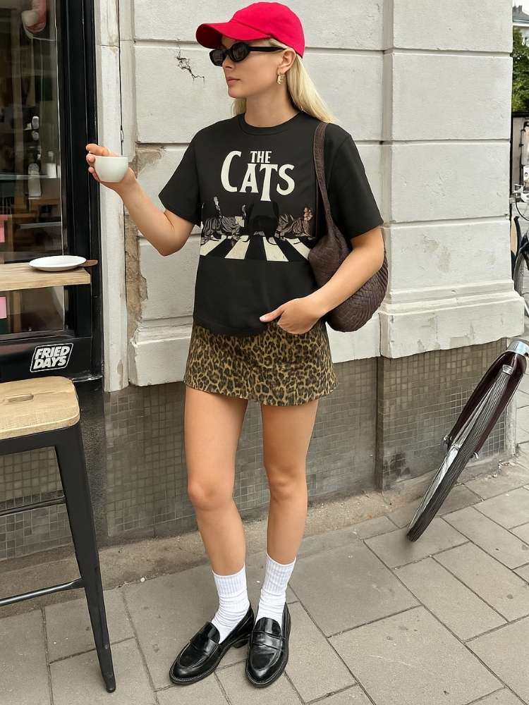 Foto de mulher usando boné vermelho camiseta preta da ALIZI, minissaia de oncinha, meia branca e mocassim de verniz preto