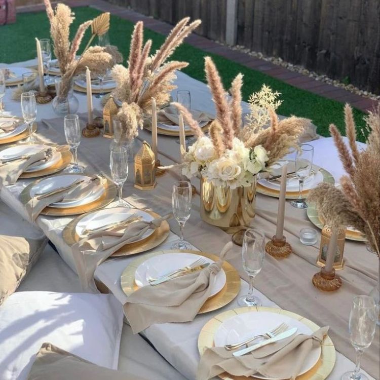 Mesa posta ao ar livre com pratos, guardanapo de tecido, almofadas no chão, toalha de mesa e arranjo desidratado, tudo em bege