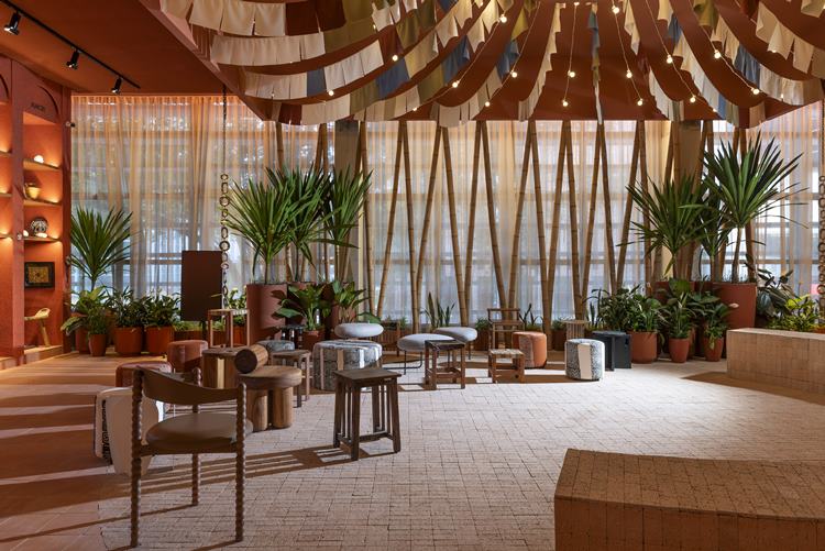 A imagem mostra um interior moderno e estiloso, com decoração tropical. Inclui cadeiras e mesas contemporâneas, plantas verdes, iluminação suave e teto com fitas de madeira curvas.

