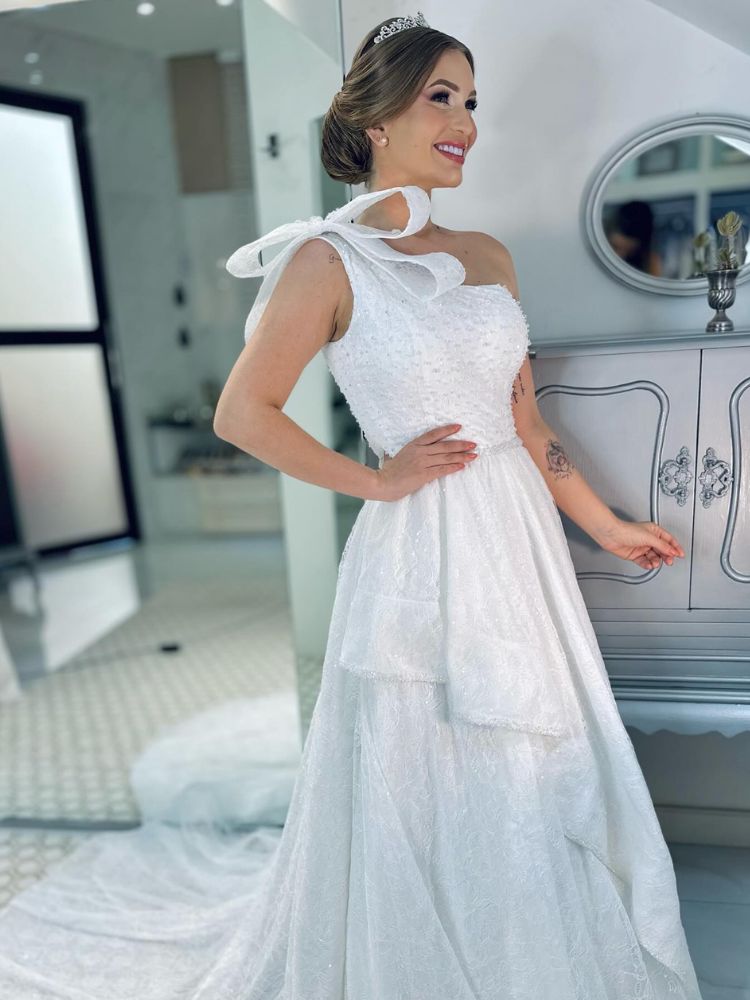 Mulher de pele clara usando vestido de noiva branco com laço grande no ombro, babados e brilho 