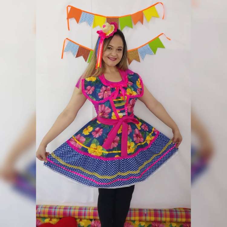Uma mulher sorridente veste um colorido vestido junino com estampa floral em tons de rosa e amarelo, e saia azul de bolinhas brancas. Ela usa um laço rosa no cabelo e exibe a saia rodada. Bandeirinhas coloridas decoram o fundo.


Fontes





