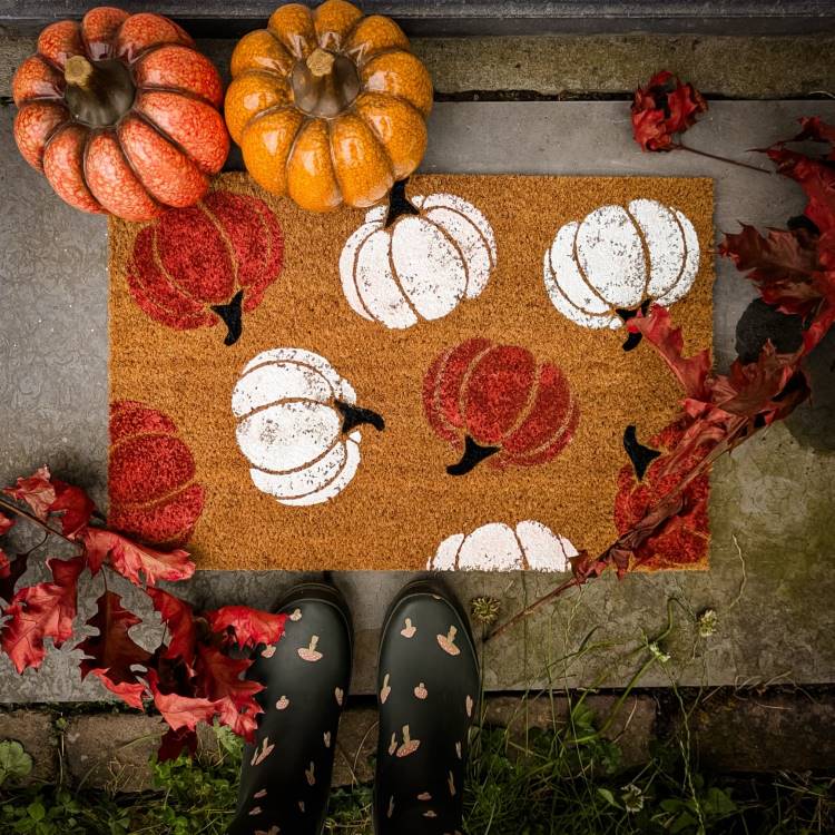 A imagem mostra um capacho decorativo com estampas de abóboras vermelhas e brancas. Ao redor, há abóboras decorativas laranja e folhas vermelhas, criando um ambiente outonal. Em primeiro plano, botas pretas com estampa de abóboras estão posicionadas sobre o capacho, completando a cena temática de Halloween.


