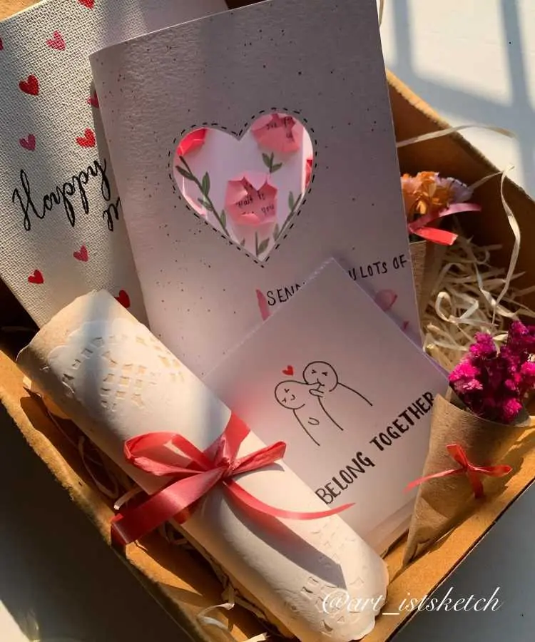 Uma charmosa caixa de presente com itens personalizados: cartão recortado em forma de coração, ilustrações minimalistas, mensagens românticas, flores secas embaladas em kraft e rolinhos decorados com laços vermelhos. Tudo disposto em palha, destacando o tema romântico e artesanal.