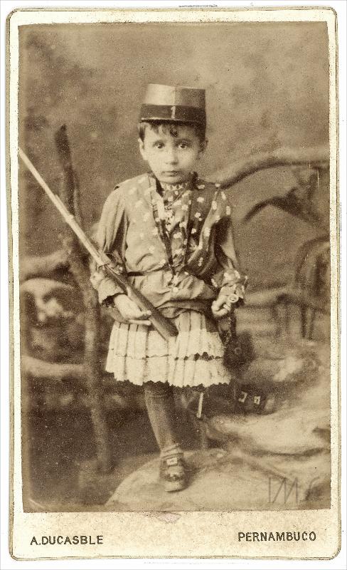 Foto em preto e branco de uma criança posando vestindo uma espécie de vestido e chapéu, com uma arma na mão Retrato de uma criança em Pernambuco por volta de 1885 vestindo uma espécie de vestido e chapéu.
