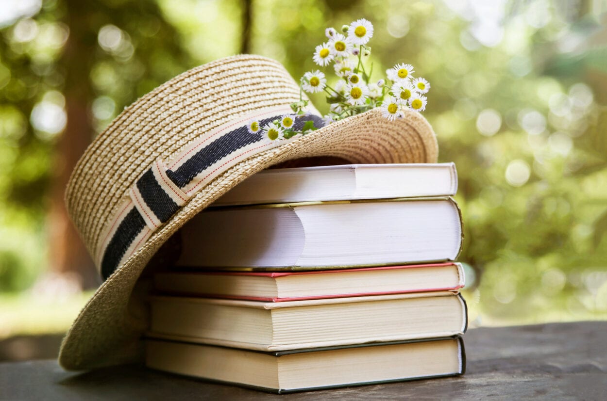 Foto de pilha de 5 livros sobre mesa marrom, com um chapéu e flores por cima