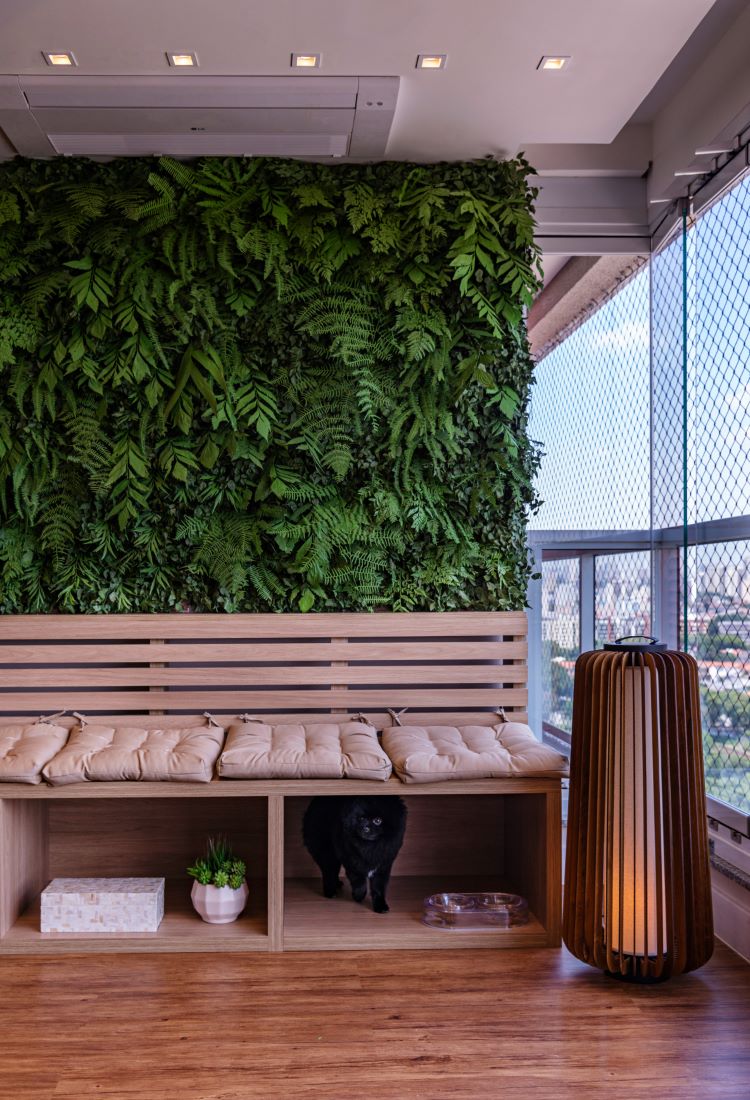 A imagem mostra uma varanda decorada com um jardim vertical exuberante, composto por diversas plantas verdes que cobrem toda a parede, trazendo frescor ao ambiente. Em frente ao jardim, há um banco de madeira com almofadas bege, que possui nichos na parte inferior, onde estão um vaso pequeno com planta, uma caixa decorativa e um espaço ocupado por um gato preto. Ao lado do banco, uma luminária de piso em madeira com luz suave complementa o ambiente. A varanda, com piso de madeira e vista para a cidade, é um excelente exemplo de como decorar apartamento com plantas, utilizando um jardim vertical para otimizar o espaço e criar uma atmosfera natural e acolhedora.


