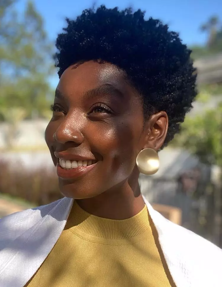 Mulher negra com cabelo crespo, make leve, usando brinco dourado e roupa amarela e branca