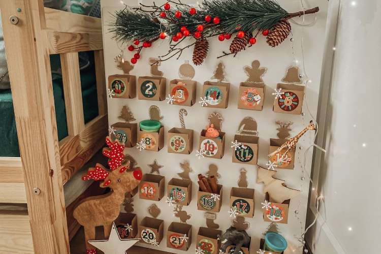 Um calendário do advento artesanal apresenta 25 caixas numeradas em uma estrutura de madeira. A decoração inclui pinhas, ramos, e bagas vermelhas no topo. Há um alce decorativo em frente, complementando o clima natalino e acolhedor do ambiente.


