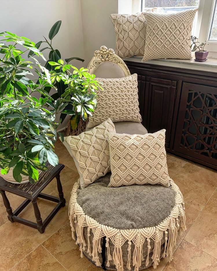 ambiente interno, possivelmente uma sala de estar, iluminado pela luz natural que entra pela janela ao fundo. Há uma cadeira ornamentada e confortável com um assento cinza e franjas penduradas na base. Três almofadas decorativas com padrões geométricos adornam a cadeira. Ao lado da cadeira, há uma pequena mesa de madeira escura que sustenta uma planta verde exuberante em um vaso. Outra planta maior está visível à esquerda da imagem. Um móvel escuro com detalhes entalhados está posicionado sob a janela.