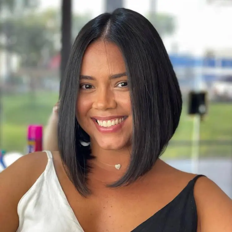 Uma mulher com um sorriso largo e cabelos pretos lisos em corte bob, divididos ao meio. Ela veste uma blusa bicolor preta e branca, e usa um colar delicado com um pingente de coração.