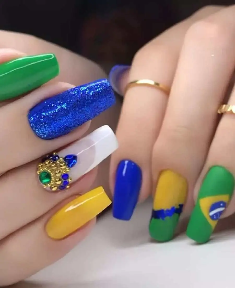 Imagem de mão com unhas decoradas para a Copa do Mundo 2022. Cores usadas, verde, amarelo, azul e branco. Polegar e indicador, azul. Médio, amarelo e verde com azul glitter entre eles. Anelar, bandeira do Brasil.