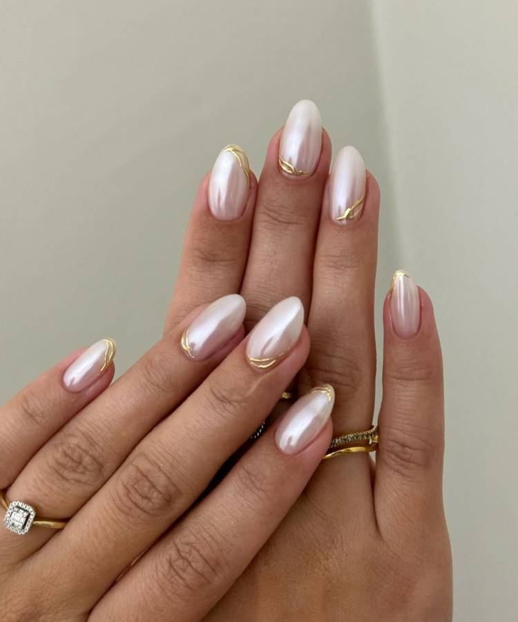 Mãos femininas com unhas em formato oval, decoradas com esmalte perolado e detalhes dourados minimalistas. Anéis delicados, incluindo um com pedra central, complementam o visual elegante. O fundo neutro realça a sofisticação e o brilho das unhas.





