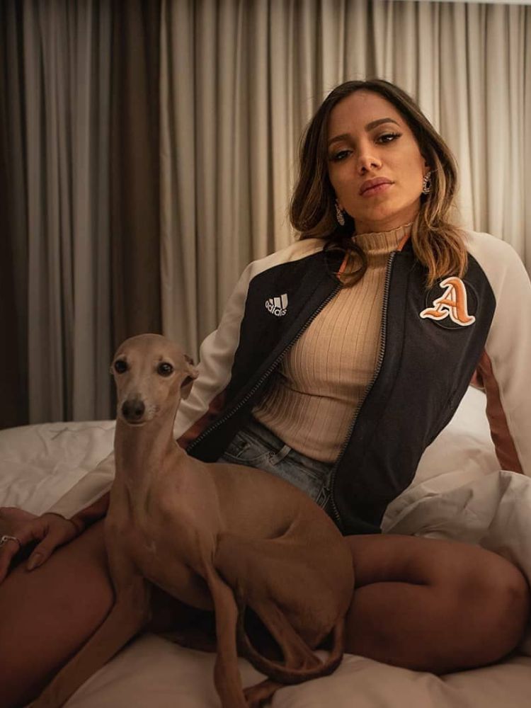 Anitta na cama usando blusa branca, shorts jeans e moletom preto, ao lado de Plínio, seu galgo italiano