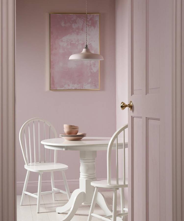 A imagem mostra um ambiente delicado com paredes rosa claro, uma mesa redonda branca e cadeiras combinando. Um quadro abstrato rosa e luminária pendente completam o espaço. Dicas de decoração para quem tem TDAH incluem cores suaves e organização visual.