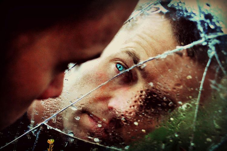 Foto de homem moreno de olhos azuis olhando sua autoimagem em um espelho quebrado