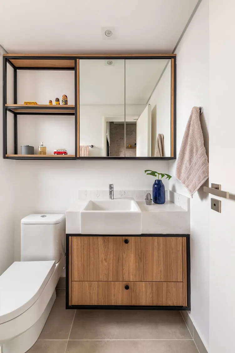 Banheiro do quarto do irmão, com pia, vaso sanitário e armário suspenso com espelho e repartições decoradas com perfume, mini carros e outros