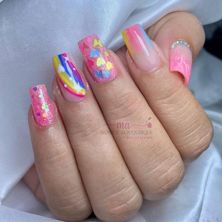 Mão com unhas arco-íris LGBTQIA+: Polegar: nude com pink e pedrinhas na cutícula Indicador: Médio: nude com arco-íris mesclado Anelar: rosa com glitter e adesivos de borboleta e coração e bolinhas nude com arco-íris e estrelas e corações Mínimo rosa com glitter 