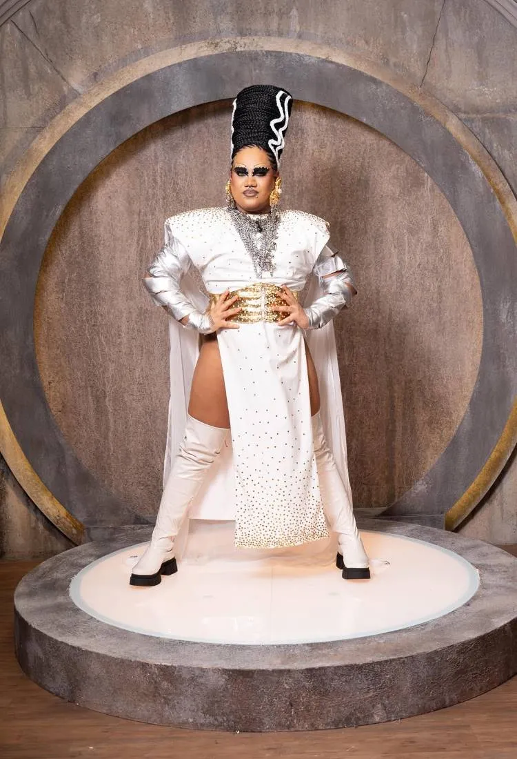 Uma pessoa vestida de **Noiva de Frankenstein**, com maquiagem pesada, uma peruca alta e preto e branca, um vestido branco e prateado com fendas e botas brancas de cano alto, posando em um palco circular.