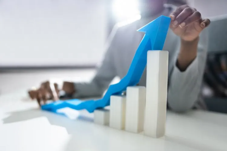 Pessoa ajustando uma seta azul sobre pilares brancos crescentes, representando crescimento, progresso e superação no contexto de negócios ou economia. Fundo desfocado com iluminação suave, destacando o movimento ascendente e a transformação positiva.
