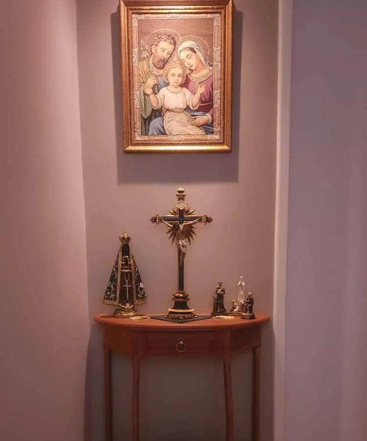 Esta imagem mostra um oratório em casa, localizado em um canto de parede. Há uma mesa de madeira com um crucifixo centralizado. À esquerda, uma imagem de Nossa Senhora Aparecida, e à direita, pequenas estátuas de santos. Acima, na parede, há um quadro com uma imagem da Sagrada Família emoldurada.