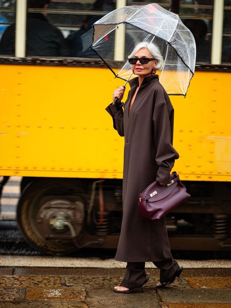 Influenciadora Grece Ghanem usando conjunto de alfaiataria marrom com sapato preto e segurando guarda-chuva e bolsa chocolate