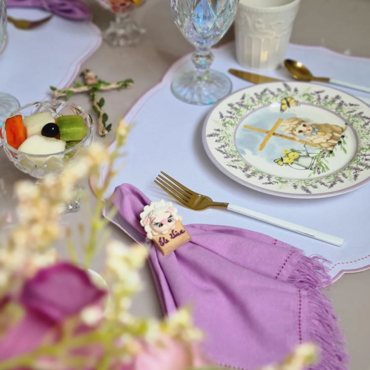 Mesa decorada com detalhes delicados, incluindo guardanapo lilás com prendedor personalizado "Ele Vive", prato com ilustração religiosa de cruz e cordeiro, talheres dourados, taças cristalinas e taça de sobremesa com frutas. Flores completam a composição elegante.