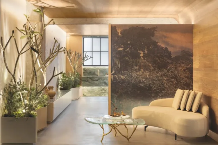 Hall de banheiro com sofá bege, mesa de centro de vidro, plantas, painel de paisagem e pias ao fundo 