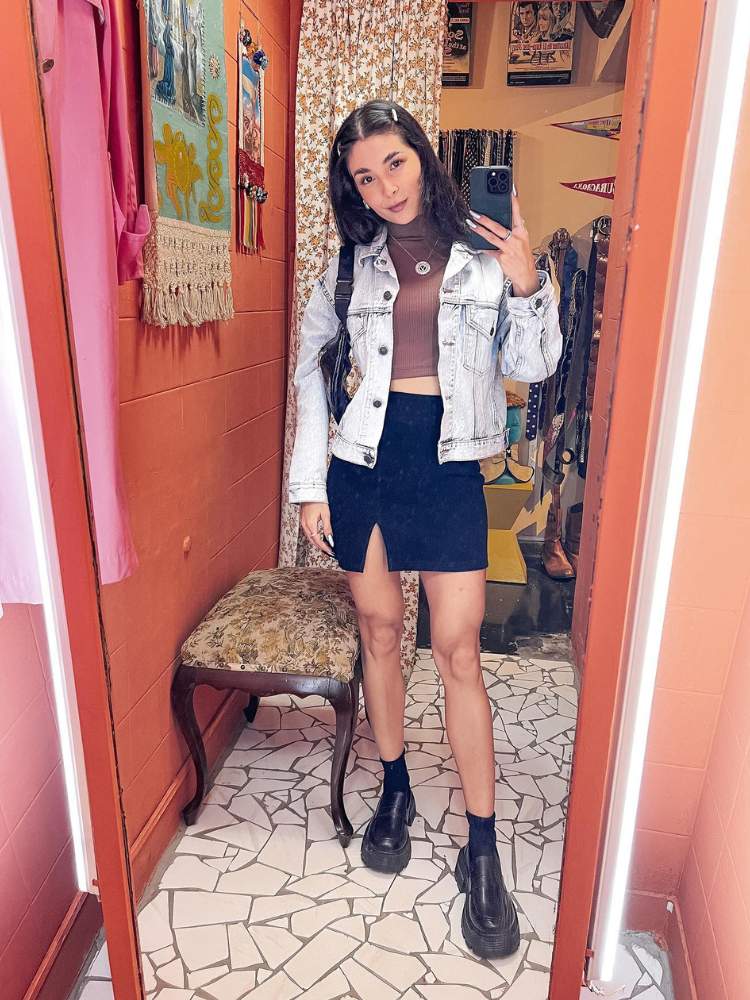 Victória de cropped marrom, saia preta, jaqueta jeans, tirando foto em espelho de provador 