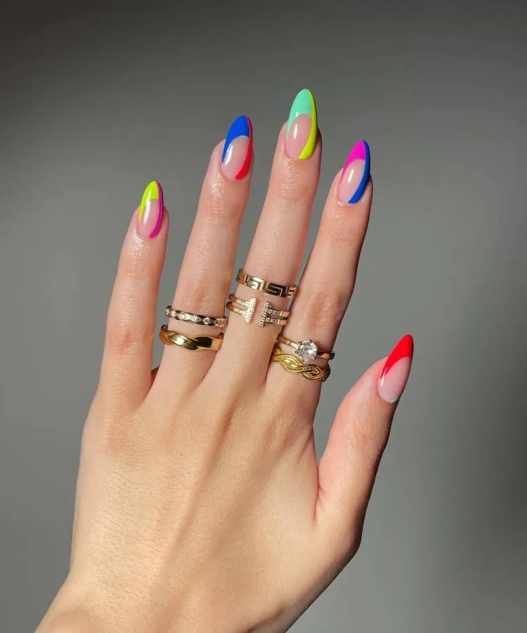 A imagem mostra um close-up de uma mão com os dedos estendidos e a palma voltada para o observador. Cada unha está pintada com um design multicolorido e brilhante, apresentando uma combinação de cores vibrantes como amarelo, rosa, verde, azul e vermelho. As unhas são longas e têm formato pontiagudo, conhecido como unhas stiletto. A mão está adornada com vários anéis em diferentes dedos; esses anéis variam em design, alguns sendo simples e outros com detalhes intrincados ou pedras embutidas. O fundo é neutro e discreto, destacando as cores vivas da arte nas unhas.