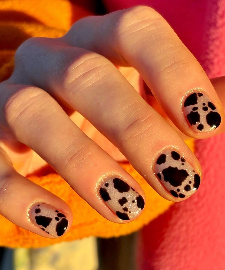 Mão com estilos de unhas curtas, decoradas em base nude brilhante e manchas pretas inspiradas em um padrão animal. A luz do sol realça os detalhes. Fundo com tecido em tons quentes, criando um visual moderno e ousado.