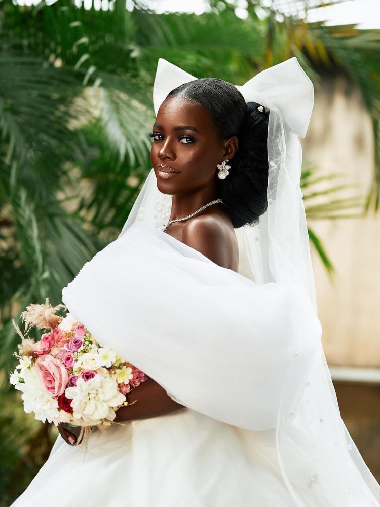 foto de mulher negra com vestido de noiva, buquê de flores rosa, brinco de flor, coque baixo e grande laço no véu