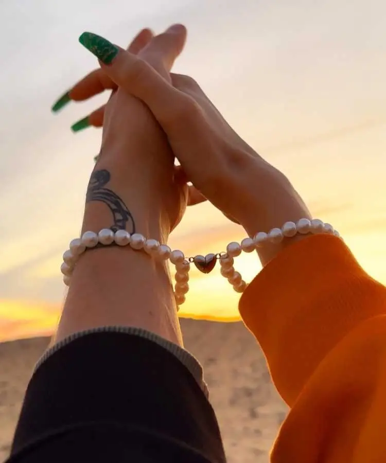 Duas mãos entrelaçadas ao pôr do sol, cada uma usando uma pulseira de pérolas conectadas por um pingente em forma de coração. Uma cena romântica com destaque para união, amor e detalhes delicados em contraste com o céu dourado.
