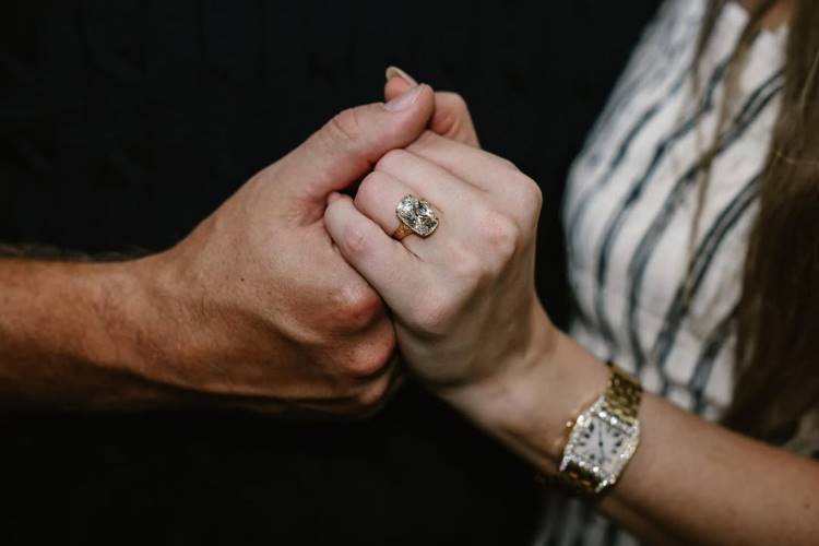 Duas mãos dadas, uma masculina e uma feminina. Na mão da mulher, um anel de noivado com uma grande pedra retangular brilha. Ela também usa um relógio de pulso dourado. O foco é no anel.