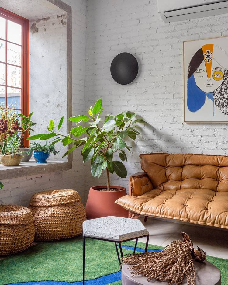 A imagem mostra uma sala de estar aconchegante e bem iluminada com decoração de verão. Há um sofá marrom de couro. Na frente do sofá, há duas caixas organizadoras redondas feitos de material trançado natural. Uma mesa lateral pequena com tampo em mármore está ao lado do sofá. Uma planta verde exuberante está em um vaso terracota no chão, adicionando um toque de natureza à sala. Outras plantas menores estão posicionadas na janela. Na parede ao lado do sofá, há uma obra de arte colorida retratando o rosto estilizado de uma pessoa. A sala tem paredes de tijolos brancos que dão um toque rústico e moderno, e uma janela grande com moldura vermelha que permite a entrada de muita luz natural. A decoração tem um toque de verão, com cores vivas e plantas que trazem um pouco da natureza para dentro de casa.

