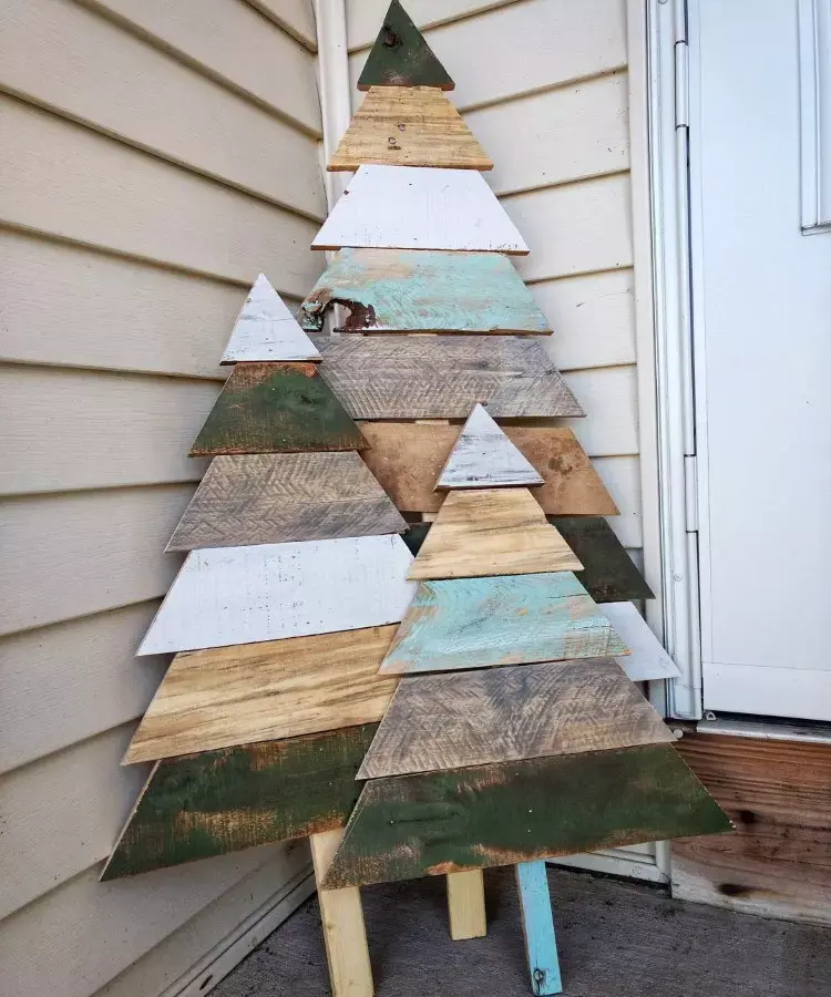 Árvores de Natal feitas de madeira reciclada, com triângulos sobrepostos em tons de verde, branco e marrom. Estão encostadas em uma parede de madeira clara, ao lado de uma porta branca. O design rústico cria um visual artesanal e natural.