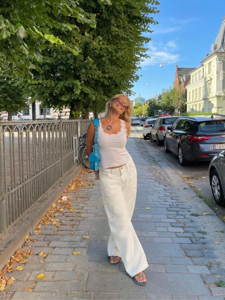 mulher usando regata branca com calça jeans branca, bolsa azul, óculos de sol e chinelo