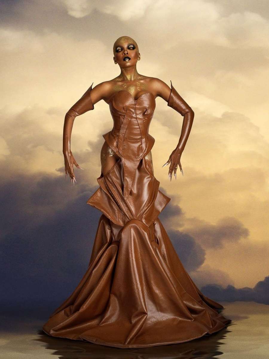Imagem em fundo com nuvens e Thelma Assis no centro. Ela veste fantasia de cobra para o Halloween Sephora. A peça é um vestido longo de cor marrom com luvas de unhas pontudas na mesma cor.