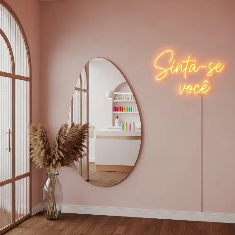 Uma luz de neon laranja com a frase "Sinta-se você" em uma parede rosa. Abaixo, um espelho oval reflete uma bancada branca com potes coloridos e um vaso de vidro com hastes secas de pluma.







