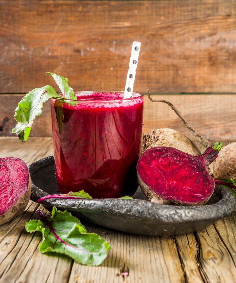 Um close-up de um copo de suco de beterraba vermelho-escuro com um canudo. Folhas e raízes de beterraba frescas e inteiras estão em uma bandeja escura ao lado do copo, tudo sobre uma mesa de madeira rústica.