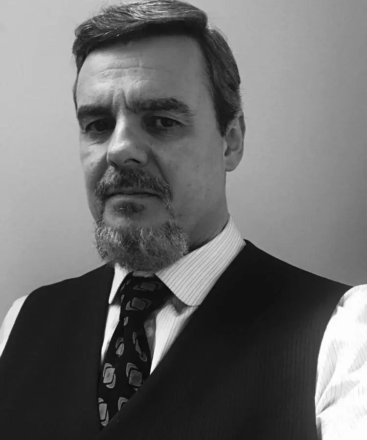 Homem de cabelos curtos e barba cheia, vestindo colete escuro, camisa listrada e gravata estampada. Fotografia em preto e branco com fundo neutro. Possível membro do elenco de "Vale Tudo" hoje reconhecido por sua elegância e expressão séria.