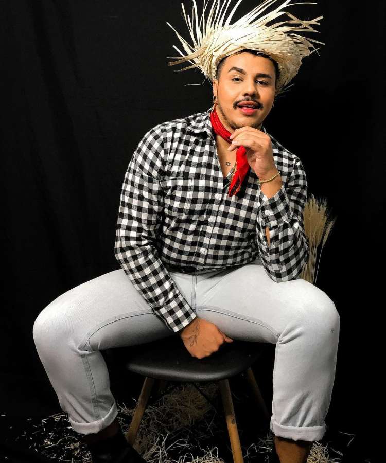 Homem com look de festa junina: camisa xadrez preta e branca, calça jeans clara, lenço vermelho no pescoço e chapéu de palha