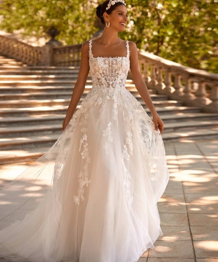 Noiva em vestido branco com corpete rendado, alças finas e saia volumosa de tule com aplicações florais, representando as tendências em vestidos de noiva 2025. O cenário ao ar livre, com escadaria e vegetação, adiciona um toque romântico.





