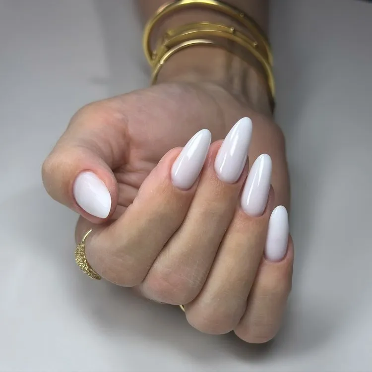 Mão feminina com unhas longas em formato amendoado, pintadas com esmalte branco brilhante, destacando elegância e simplicidade. Acessórios dourados, como pulseiras e anel minimalista, complementam o visual sofisticado. Fundo neutro reforça o foco no design delicado e moderno das unhas.