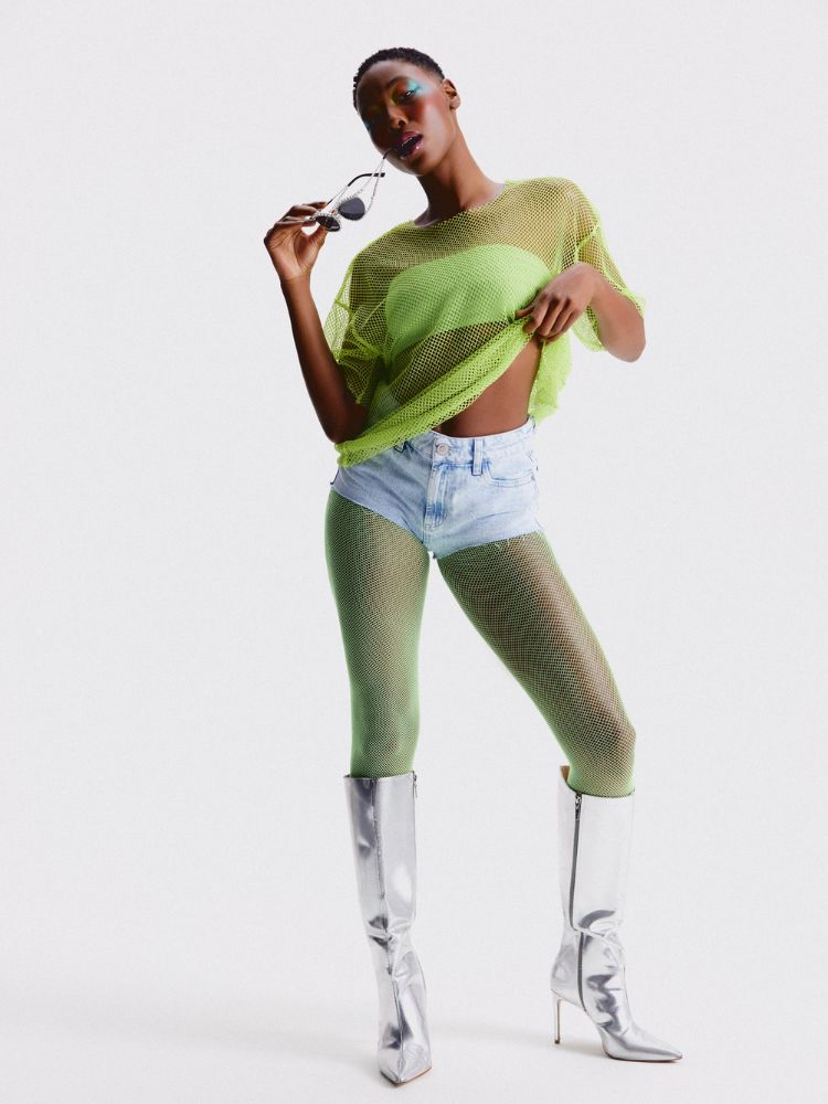Mulher de pele clara usando top verde, camiseta de tule por cima, meia calça verde, shorts jeans e bota metalizada 