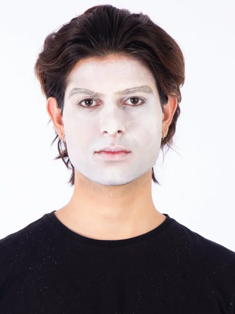 Homem de pele clara, cabelo médio castanho, usando blusa preta e pancake branco no rosto 