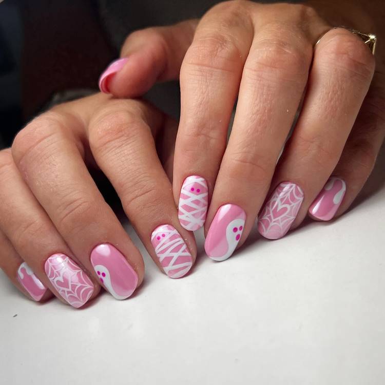A imagem mostra unhas decoradas em tons de rosa e branco, com desenhos temáticos de Halloween. Há fantasmas, teias de aranha e padrões de múmia, todos estilizados. As unhas têm formato oval e um acabamento brilhante, criando um visual divertido e criativo. Ideal para quem deseja um toque festivo e temático nas unhas.