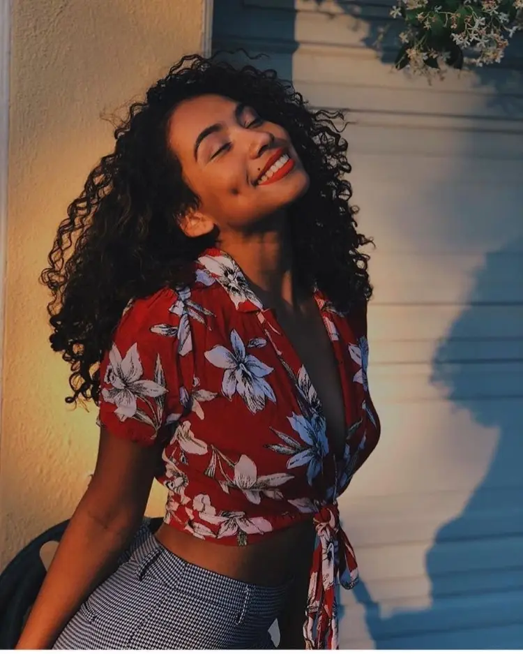 Frase para foto pensativa mulher negra sorrindo com cabelo cacheado, usando batom vermelho, cropped vermelho com estampa floral e calça quadriculada preto e branco Frase para foto pensativa mulher negra