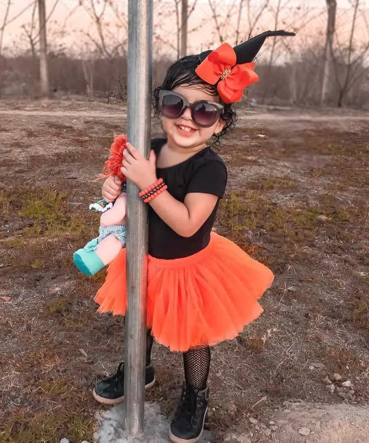 Menina com fantasia de bruxa infantil. Cores predominantes: preto e abóbora. Usa: body, saia de tule, meia arrastão, tênis, pulseiras, tiara de chapéu pontudo com laço, óculos de sol, segura boneca