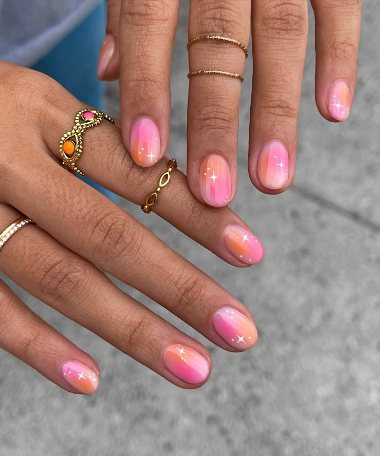 Mãos com unhas decoradas em degradê de rosa e laranja, finalizadas com pequenos brilhos brancos desenhados. Os dedos estão adornados com anéis delicados de ouro e pedras coloridas. Fundo cinza desfocado destaca o design elegante e vibrante das unhas.