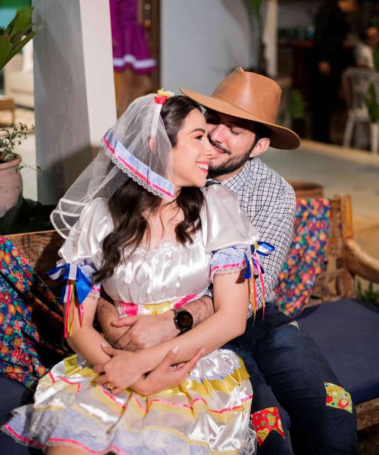 Um retrato terno de um casal com trajes típicos de Festa Junina. O homem, de camisa xadrez e chapéu de cowboy, abraça a mulher, que veste um vestido branco e amarelo com fitas e véu. Eles sentam em um banco de madeira, seus sorrisos refletindo carinho em um cenário festivo e decorado.